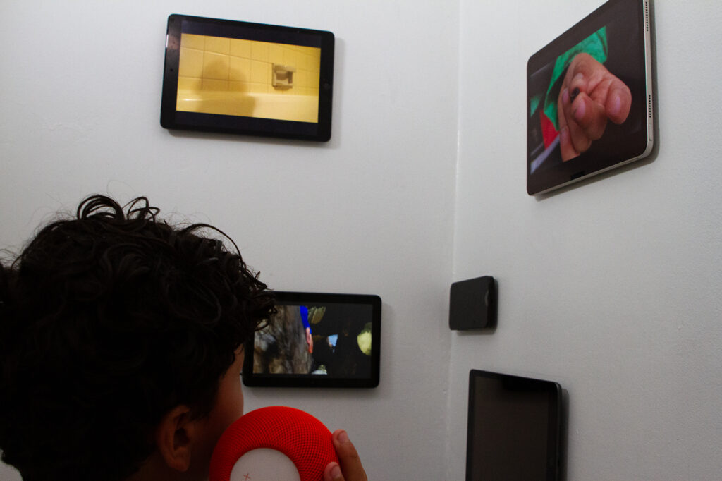 Nesting — installation detail with viewer Viewer holding a orange Bluetooth speaker while watching several screens from Nesting installed across two walls.