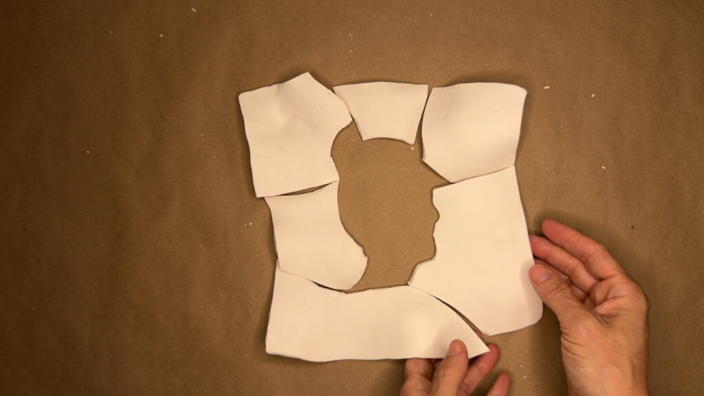 Con la esperanza de empatar rostros con nombres, process documentation, 2026 Two hands hold together broken white porcelain puzzle pieces on a brown kraft paper surface, revealing a silhouette-shaped void at the center where pieces are missing or do not align.