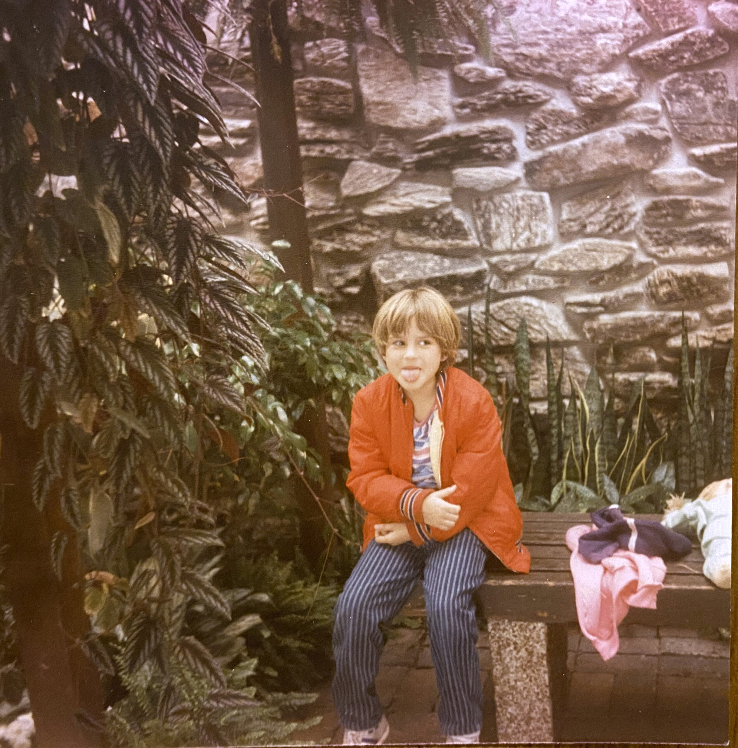 Nicole, 1984 A young child sits on a wooden bench in a stone-walled indoor garden, sticking her tongue out at the camera. She wears a red windbreaker over a striped shirt and striped pants. A doll lies on the bench beside her.